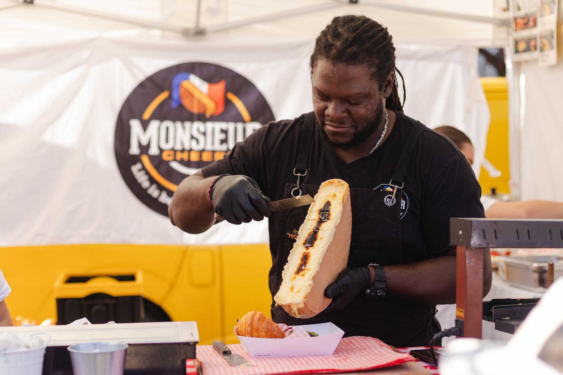 Chef scraping melted cheese onto food at outdoor booth, with sign in background.