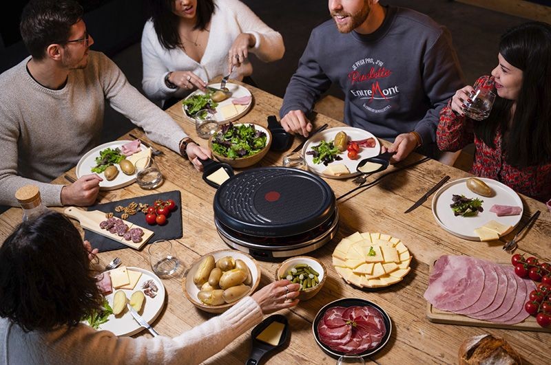Group of people enjoying a meal with raclette grill and various foods on a wooden table.